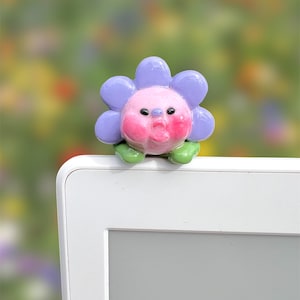 May include: A whimsical, handcrafted flower-shaped figurine with a smiling face. The flower has lavender petals, a pink face with rosy cheeks, and green leaves. It is perched on a white electronic device, set against a blurred floral background.