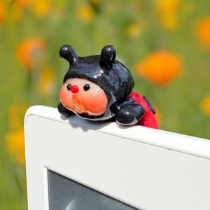May include: A small, glossy glass figurine shaped like a ladybird with a black head and body, a bright orange face, and a tiny red nose. The figurine features two black antennae and a red wing section, perched on the top edge of a white screen.