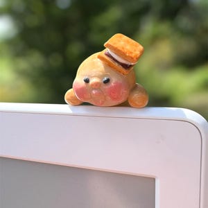 May include: A whimsical, light-brown figurine with rosy cheeks and a small, square s'more on its head. The figurine has small black eyes and is resting on a white surface. The background is a blurred green, suggesting an outdoor setting.