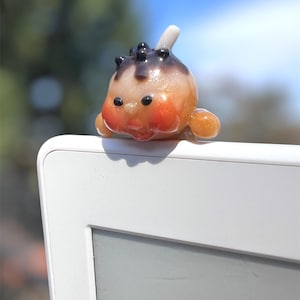 May include: A whimsical, orange and brown glass figurine resembling a small creature, with black accents and a white protrusion. The figurine is perched on the edge of a white electronic device, set against a blurred background of green and blue.