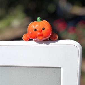 May include: A small, orange pumpkin figurine with a green stem and a face. The pumpkin has small arms and is resting on a white surface. The background is blurred with green and red hues.