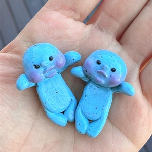 May include: Two small blue ceramic figurines with speckled paint and purple accents. The figurines have a human-like shape with round heads and small limbs.
