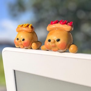May include: Two quirky hot dog figures with smiling faces and arms. One has yellow mustard, the other red relish. The figures are light brown with rosy cheeks, and sit on a white surface.