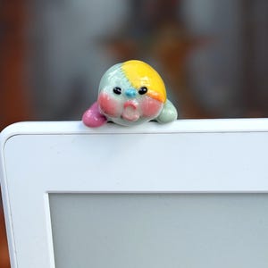 以下が含まれることがあります： 驚いた表情をした丸い生き物の形をした小さな光沢のある陶器の置物で、淡い緑色の体、頭の鮮やかな黄色の斑点、ピンクの頬、小さな紫色の手足が特徴で、白いフレームの端に置かれています。