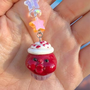 May include: A handmade cupcake charm keychain with a red body, white frosting with red sprinkles, and a purple base. The charm is connected to a silver chain with iridescent star and bead accents.
