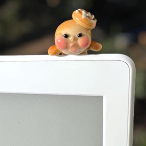 May include: A whimsical ceramic figurine of a character with rosy cheeks and a cinnamon roll on its head. The figurine is tan with brown and white accents. It is perched on the edge of a white electronic device.