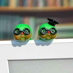 May include: Two small, round, bright green glass figurines with prominent cheeks, red noses, and black-rimmed glasses. One figurine wears a black graduation cap on its head. Both figures have a smooth, glossy finish and are positioned side by side.