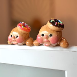 May include: Two small ceramic figurines with round faces and rosy cheeks, each wearing a donut hat. One donut is pink with colorful sprinkles, the other is brown with chocolate frosting and sprinkles.