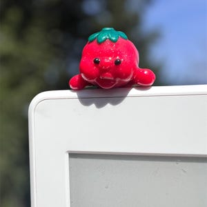 May include: A red and green strawberry-shaped figurine with black eyes and a white background.