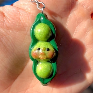 May include: A green pea pod keychain with three peas. The middle pea has a face with rosy cheeks and a small mouth. The keychain is attached to a silver chain.