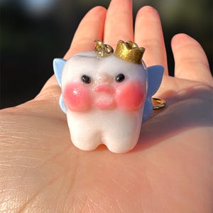 May include: A whimsical tooth-shaped charm with a gold crown, rosy cheeks, and blue fairy wings. The charm has black eyes and a small mouth. It is a unique accessory.