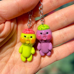 May include: Two small, glossy, cartoon-style character charms attached to silver-toned metal keychains. One charm is lime green with a pink brain-like top, and the other is purple with a green brain-like top. Both have round faces.