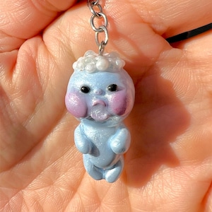 May include: A handmade keychain featuring a small, blue figurine with a baby-like appearance. The figurine has rosy cheeks, black eyes, and a white, pearl-like headpiece. The keychain is attached to a silver chain.