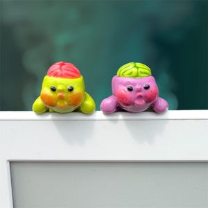 May include: Two small, rounded figurines perched on a white edge. The left figurine is lime green with a bright red brain-like top. The right figurine is purple with a light green brain-like top. Both have wide eyes and open, surprised mouths.