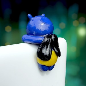 May include: A small, glossy figurine shaped like a character with a rounded blue head and body, featuring black wings and a yellow midsection. The figure is designed to hook over a thin edge, with its arms resting on top and its body hanging down.