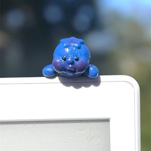 May include: A charming, blue blueberry-shaped ornament with a shiny surface. The figurine features tiny arms and a face with black eyes and pink cheeks. It is positioned on the edge of a white surface, with a blurred green and blue backdrop.
