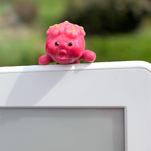 May include: A pink figurine with a flower crown and a small, round face. The figurine is made of polymer clay and has a glossy finish.