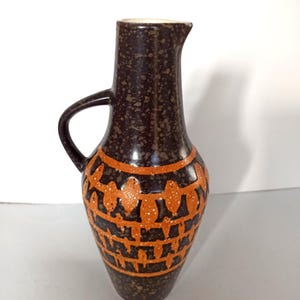 May include: A brown ceramic pitcher with a handle and spout. The pitcher has a speckled finish and is decorated with orange geometric patterns. The interior of the pitcher is white.