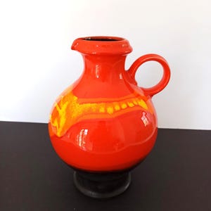 Könnte beinhalten: Eine leuchtend orangefarbene Keramikkrug mit rundem Körper, Henkel und Ausguss. Der Krug hat ein gelb-oranges Muster in der Mitte und steht auf einem schwarzen Sockel. Ein dekoratives Objekt für das Zuhause, ca. 20 cm hoch.