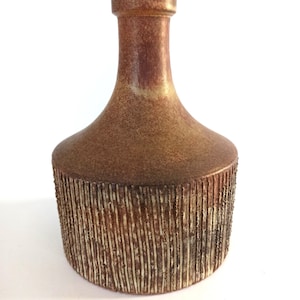 Może przedstawiać: Brązowy ceramiczny wazon z teksturowanym, pionowym wzorem na dolnej części. Wazon ma zaokrąglony korpus i smukłą szyję, z nakrapianą, ziemistą paletą kolorów.