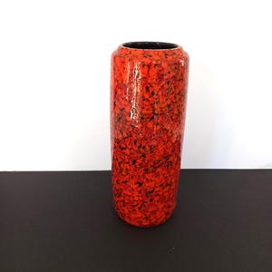 Op de afbeelding: Een cilindervormige vaas met een gespikkeld rood en zwart patroon. De vaas heeft een glanzende afwerking en een zwart interieur. Het ontwerp doet denken aan vulkanisch gesteente. De vaas is een decoratief item.