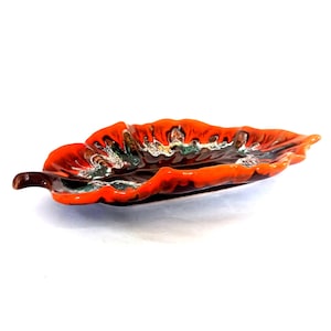 Puede incluir: Un plato de cerámica con forma de hoja y un vibrante esmalte naranja. El plato presenta un tallo marrón oscuro y detalles interiores verdes y blancos. Esta pieza decorativa es ideal para servir aperitivos o como un acento único para el hogar.