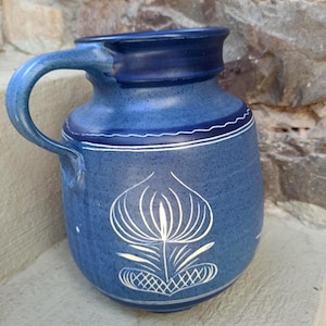 Könnte beinhalten: Ein blauer Keramikkrug mit gesprenkelter Oberfläche und einem einzelnen Henkel. Der Körper zeigt ein weißes, handgemaltes Blumenmotiv und eine dünne Wellenlinie um den oberen Bereich. Der Rand ist in einem dunkleren Blauton gehalten und abgesetzt.