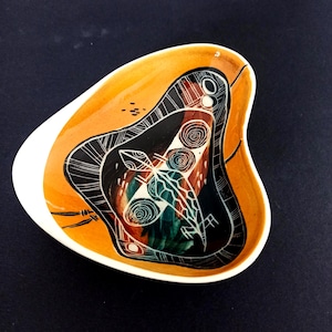 Op de afbeelding: Een asymmetrische keramische schaal met een witte buitenkant en een feloranje binnenkant. Het midden bevat een donker, abstract grafisch ontwerp met witte lijntekeningen, waaronder spiraalmotieven en een centrale insectachtige figuur.