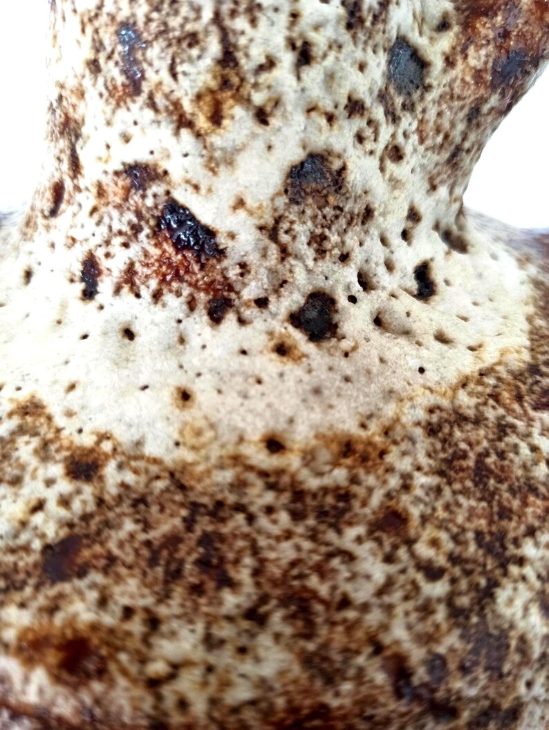 May include: Close-up of a ceramic pitcher's textured surface. The pitcher has a speckled, porous design with shades of brown and cream. Darker brown spots and small holes create a unique, rustic appearance. The pitcher's handle is visible in the upper right corner.