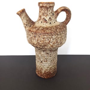 Può includere: Brocca in ceramica marrone e beige strutturata, con beccuccio e manico. La brocca ha un corpo bulboso e una base cilindrica. La superficie ha un aspetto maculato, con macchie marroni più scure. Il beccuccio è angolato e il manico è un semplice anello.