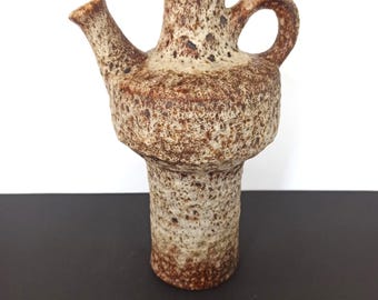 May include: A textured, brown and beige ceramic jug with a spout and handle. The jug has a bulbous body and a cylindrical base. The surface has a speckled appearance, with darker brown spots. The spout is angled, and the handle is a simple loop.