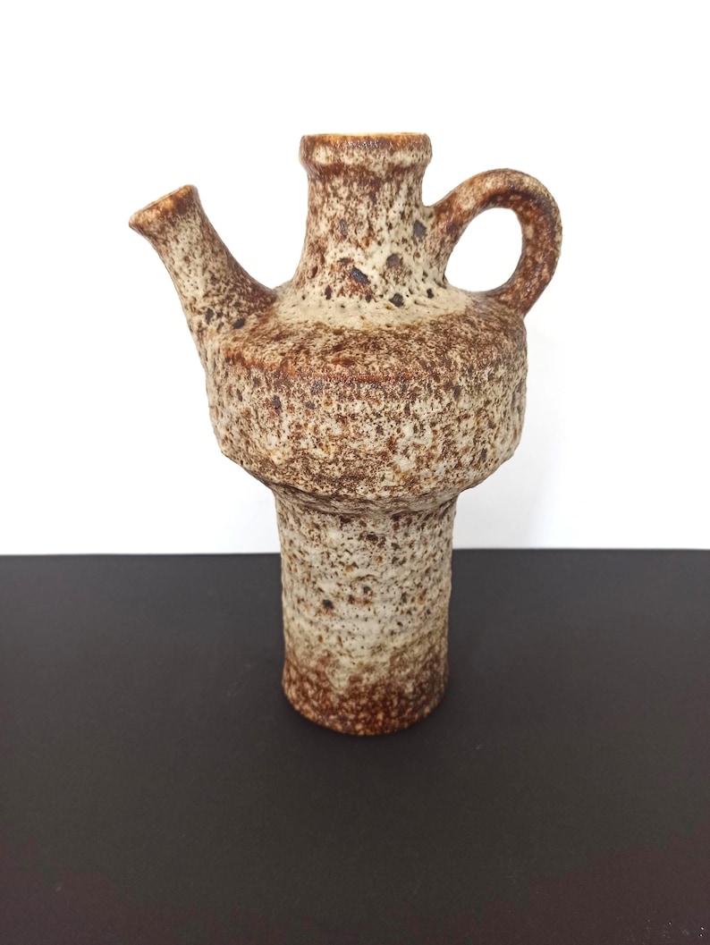 May include: A textured, brown and beige ceramic pitcher with a spout and handle. The pitcher has a bulbous body and a cylindrical base. The surface has a speckled appearance, with darker brown spots. The spout is angled, and the handle is a simple loop.