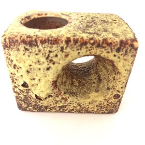 May include: A rectangular ceramic object with a textured, speckled yellow and brown glaze. The piece features a circular opening on the top surface and a large, arched cutout through the center, creating a hollow, sculptural form with a rough finish.