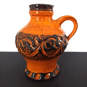 Könnte beinhalten: Orangefarbener Keramikkrug mit Henkel und strukturierter Oberfläche. Der Krug hat einen dunkelbraunen Rand und Boden, mit einem Band aus schwarzen floralen Designs in der Mitte. Ein kleines, dekoratives Stück.