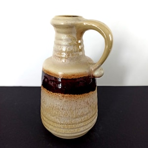 May include: A small, ceramic pitcher with a handle. The pitcher is beige with a dark brown horizontal stripe around the middle. The handle and top are a lighter beige color. The pitcher has a textured surface.