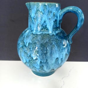 May include: A ceramic pitcher featuring a rounded body and a curved handle. The surface is finished with a mottled glaze in shades of bright turquoise, deep blue, and dark teal, creating a layered, dripped effect across the entire exterior.
