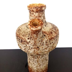May include: A textured ceramic vase with a unique design. The vase has a light beige base color with brown speckles and accents. It features a cylindrical neck and a smaller spout on the side. The vase is approximately 7 inches tall.