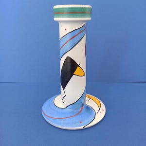 Może przedstawiać: Biały ceramiczny świecznik o fantazyjnym wzorze. Świecznik ma cylindryczny korpus z zielono-czerwonym paskiem na górze i okrągłą podstawą. Korpus zdobią niebieskie, czarne i żółte abstrakcyjne kształty i linie.