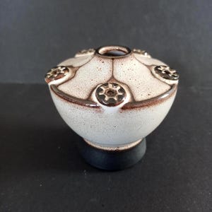 Peut inclure: Un pique-fleurs en céramique avec un corps arrondi blanc cassé, des taches brunes et une base brun foncé. La partie supérieure présente une ouverture centrale entourée de cinq petits orifices circulaires, chacun orné d'un bord en forme d'étoile.