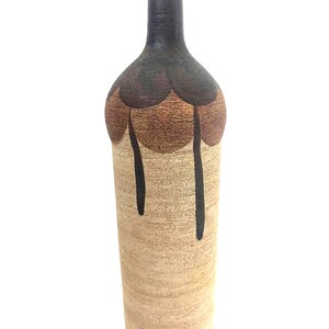 Op de afbeelding: Een hoge keramische fles met een smalle hals, voorzien van een getextureerde beige basis en een donkerbruin bovengedeelte. De schouder van de fles is versierd met een geschulpt patroon in bruintinten, met donkere glazuurdruppels die naar beneden lopen.