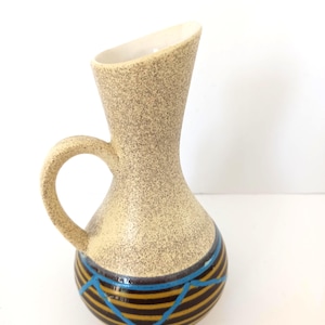 Op de afbeelding: Een beige keramische kan met een gespikkelde afwerking en een gebogen handvat. De basis heeft horizontale zwarte en gouden strepen met blauwe geometrische accenten. De binnenkant is wit.