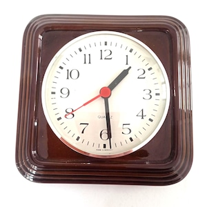 May include: A square brown ceramic wall clock with a white circular face, black Arabic numerals, black hour and minute hands, and a red second hand. The clock face features the text QUARTZ and MADE IN GERMANY at the bottom center.