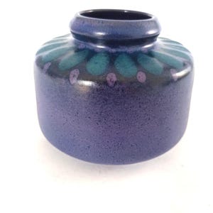May include: A small, round ceramic vase in shades of blue and teal. The vase has a textured, speckled finish and a decorative pattern of teal ovals and purple dots around the top. The opening is dark blue.