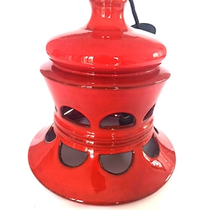 Peut inclure: Une lanterne en céramique rouge vif avec une finition brillante. La lanterne présente un dessus arrondi, un corps cylindrique avec des découpes décoratives et une base évasée. Une poignée noire est fixée au sommet. Le design est symétrique et l'esthétique générale est moderne.