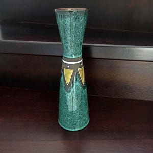 May include: A teal ceramic vase with an hourglass shape. The vase has a speckled glaze and a decorative band with gold and black geometric designs. The vase is approximately 20 cm tall.