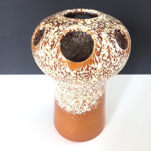 Może przedstawiać: Ceramiczny wazon w kształcie grzyba z nakrapianym brązowo-białym wzorem. Wazon ma zaokrągloną górę z kilkoma okrągłymi otworami i cylindryczną podstawą. Dolna część jest w jednolitym brązowym kolorze.