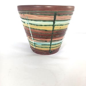 Puede incluir: Un jarrón decorativo de cerámica con una parte superior ancha y abierta y un cuerpo cónico. El jarrón presenta bandas horizontales de color, incluyendo marrón, melocotón, amarillo y turquesa, con acentos verticales verde oscuro. Decoración del hogar.