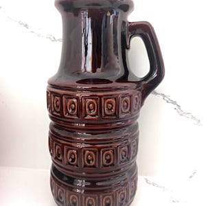 May include: A glossy, dark brown ceramic vase with a handle. The vase has a cylindrical body with a series of embossed square patterns. The top flares out slightly, and the handle is a curved, rectangular shape. The vase is likely vintage.
