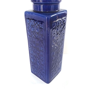 Könnte beinhalten: Eine hohe, quadratische Keramikvase mit einer glänzenden, tiefblauen Glasur. Die Außenfläche weist ein sich wiederholendes, geprägtes Gittermuster aus verschiedenen kleinen geometrischen Formen, Symbolen und Linien auf. Mit breitem, zylindrischem Hals.