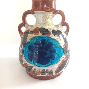 May include: A ceramic vase with two handles, featuring a cream-colored base with a crackle glaze finish. The body is decorated with a large, embossed blue flower motif surrounded by dark brown and black leaf-like patterns and a brown rim.